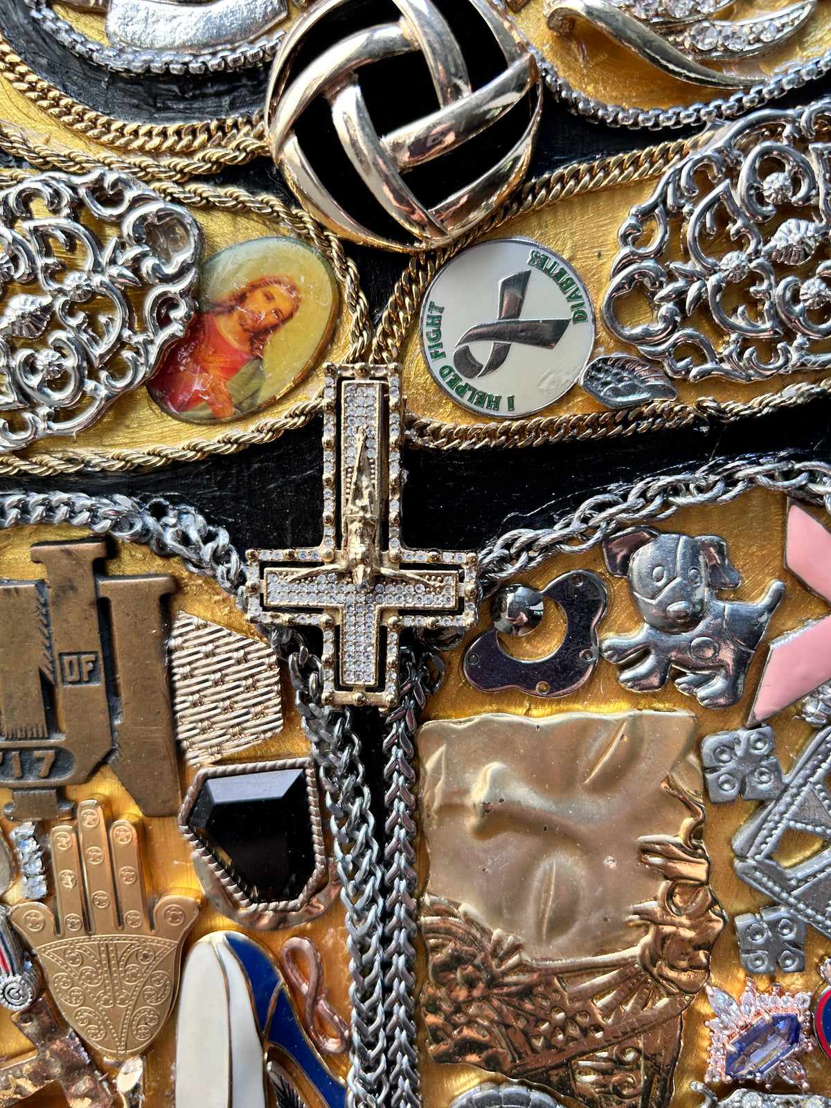 A close-up collage of assorted jewelry and religious symbols, including a large silver cross with crystals, multiple gold-toned chains, brooches, medallions, and metallic charms on a dark background.