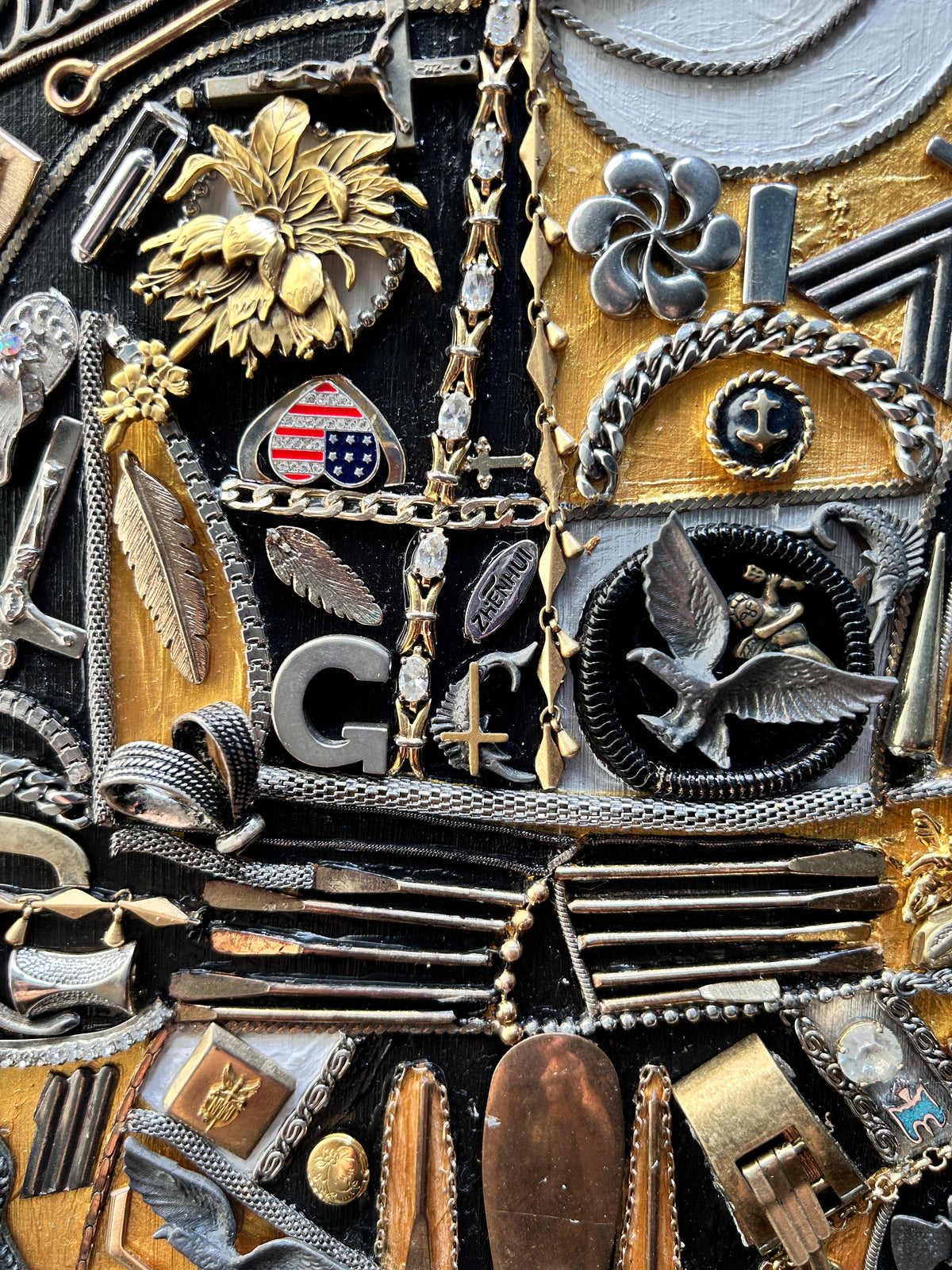A close-up of a highly detailed, textured collage artwork composed of numerous vintage jewelry elements, including silver and gold chains, brooches, rings, and charms, featuring a central letter 'G' and a bird motif.