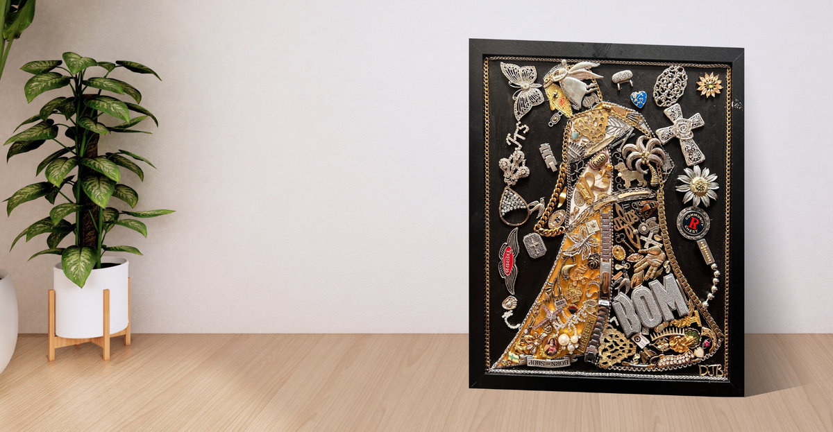 A framed decorative artwork featuring a stylized figure in ornate gold and silver garments with jewelry and floral motifs, displayed on a light wood surface against a white wall next to a potted plant.